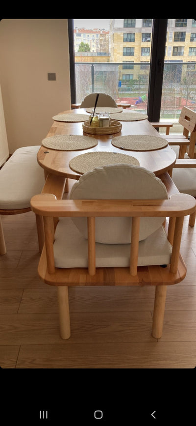 Pencere önünde yuvarlak ahşap yemek masası, beyaz sandalyeler ve ön planda ahşap kollu koltuk