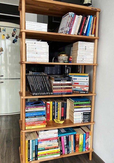 Buzdolabının yanındaki geniş ahşap kitaplık; raflarda kitaplar, CD kutuları ve dergiler