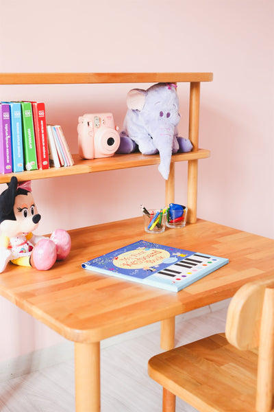 Çocuk odasında ahşap çalışma masası ve raflı kitaplık; raflarda renkli kitaplar ve peluş oyuncaklar