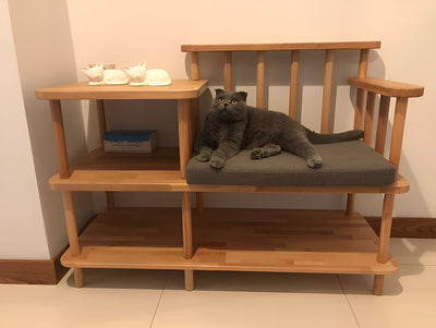 Kademeli ahşap benchin gri minderinde uzanmış gri kedi; üst tablada iki seramik kedi figürü