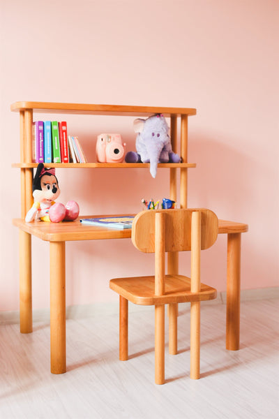 Pembe duvarlı çocuk odasında ahşap çalışma masası, üstünde raflı kitaplık ve ahşap çocuk sandalyesi