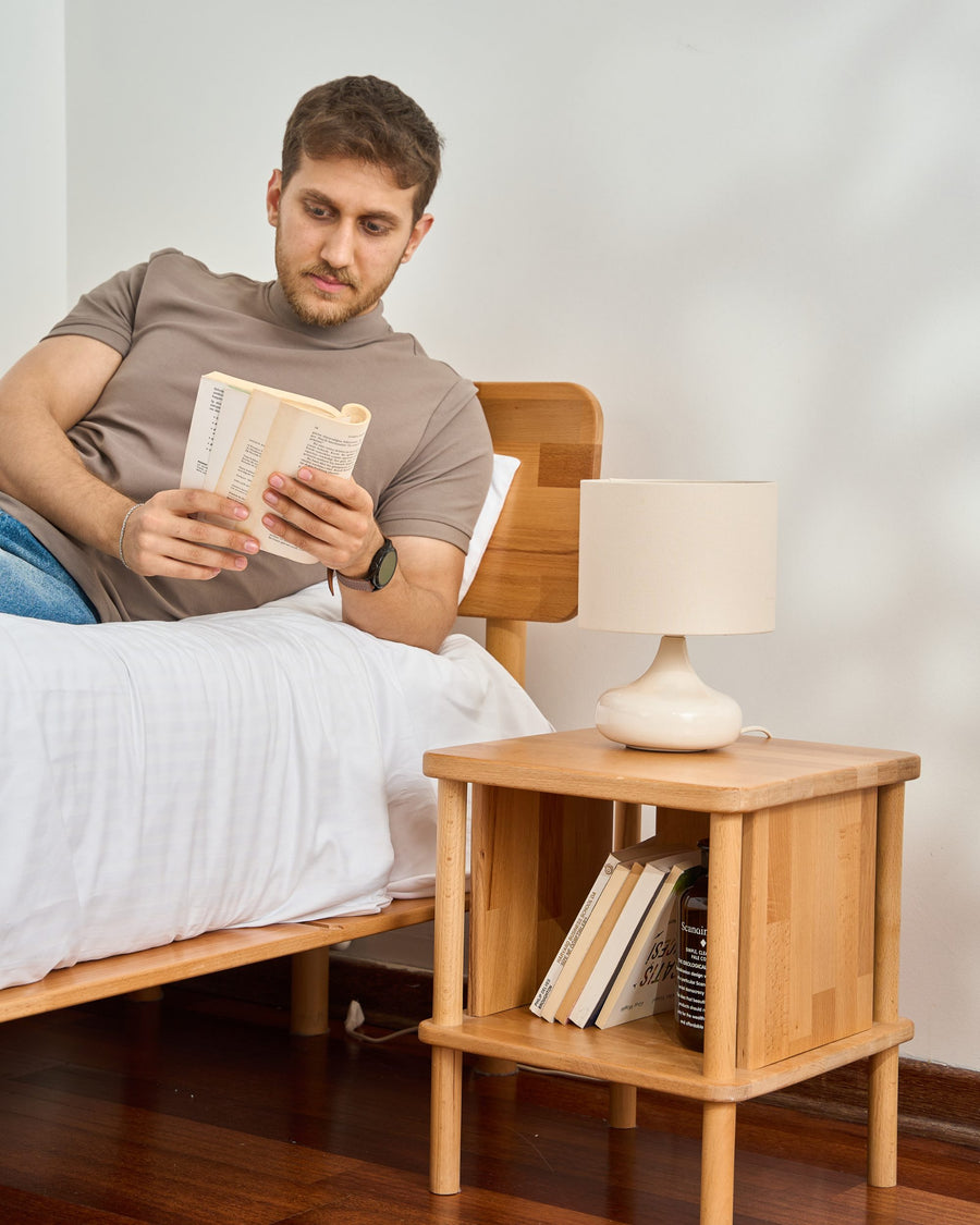 Komodin ve lamba yanında yatakta kitap okuyan kişi